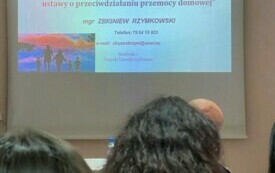 Biała plansza na kt&oacute;rej wyświetlone są czerwone oraz niebieskie litery z nazwą szkolenia oraz danymi kontaktowymi prowadzącego. Przed planszą siedzący uczestnicy szkolenia.